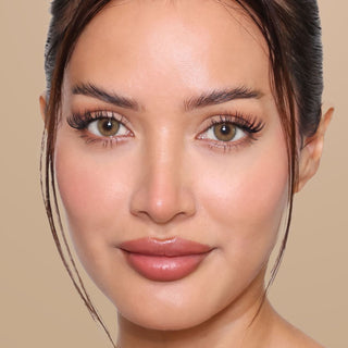 A woman with smooth, glowing skin, full lips, defined eyebrows, long lashes, and brown hair pulled back, shown against a neutral beige background.