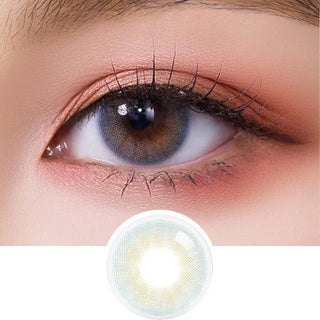 A close-up of an eye with a brown iris, enhanced with warm makeup and long lashes. Below the eye, Olola Able Blue lenses by Olola are displayed, offering a natural look for dark eyes with a subtle blue gradient pattern.