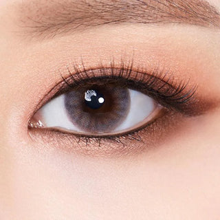 A close-up of an eye wearing Olola Amuse Grey by Olola, featuring long eyelashes, smooth skin, and soft brown eyeshadow. The iris is a striking grey shade, contrasting beautifully with natural makeup details.