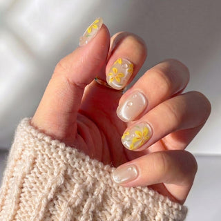 A hand with EyeCandys After Rain Press-On Nails—sheer nude base, yellow flower designs, and 3D droplets—shows off reusable nails against a creamy knit sweater sleeve.