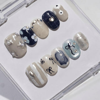 The All Tied Up Press-On Nails by EyeCandys feature handmade designs—flowers, abstract patterns, metallic accents, and zodiac-style symbols—in beige, blue, white, and silver, all displayed in a clear case.