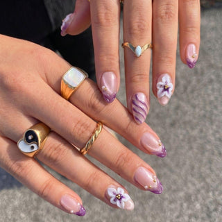 A close-up of hands wearing EyeCandys Aloha Darling Press-On Nails, featuring purple tips, white and purple floral designs, swirls, and unique gold rings—perfect for showcasing tropical nail art with intricate accents.