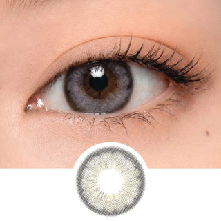A close-up of a person's eye wearing EyeCandys Pink Label Angel Grey contact lenses, featuring a gray and light yellow pattern. Below the eye is an enlarged image showing the design of these circle lenses.