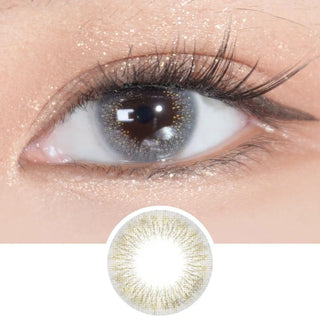 A close-up of a brown eye with long lashes wears EyeCandys Attitude Glitter Grey color contact lenses, featuring gold accents and shimmering gold eyelid makeup. An enlarged view of the EyeCandys lens design appears below the eye.