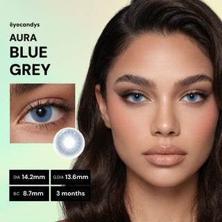 A woman with long brown hair wears EyeCandys Aura Blue Grey contact lenses by EyeCandys—DIA 14.2mm, G.DIA 13.6mm, B.C. 8.7mm, 3 months. “AURA BLUE GREY” text and a close-up of the lens on an eye are shown. Designed for brown eyes.