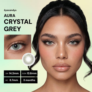 A woman with light skin and dark hair wears EyeCandys Aura Crystal Grey contact lenses with UV blocking. A close-up of her eye showcases the lens detail. Text shows "EyeCandys Aura Crystal Grey," specs, and a 3-month replacement period.