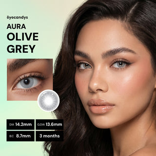 A woman with smooth, glowing skin and long dark hair wears EyeCandys Aura Olive Grey contacts. A close-up highlights her eye, lens details, and the EyeCandys Aura Olive Grey product name on the left of her face.