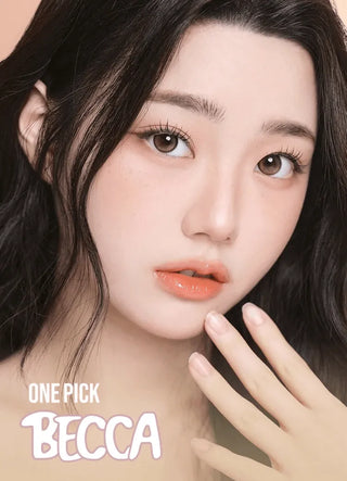 A young woman with long, dark hair and flawless skin poses with her hand near her face, wearing natural makeup and glossy peach lips. She has on i-Girl One Pick Becca Azuki Brown contacts. The text "ONE PICK BECCA" is at the bottom of the image.