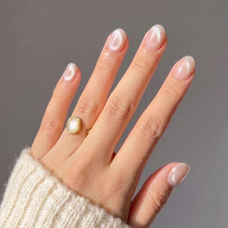 A hand with short, rounded Bare Minimum Press-On Nails by EyeCandys in pearly white, mimicking a salon gel manicure. The model wears a cream knit sweater and a gold ring with a white stone; background is plain and neutral.