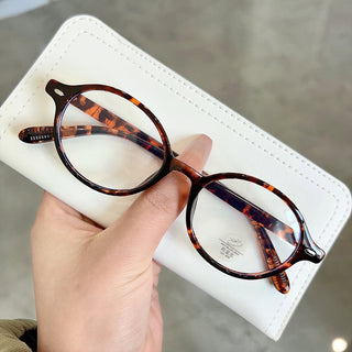 A hand holds a pair of EyeCandys Chase Round Oval Glasses with clear lenses, their round tortoiseshell frames resting on a white case, set against a smooth grayish background.