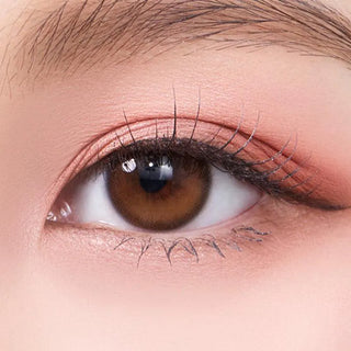 A close-up of an eye wearing Olola Dahlia Brown color contact lens by Olola, with long lashes and soft pink eyeshadow on smooth, flawless skin.