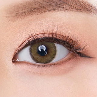 A close-up shows an eye wearing Olola Dearsome Olive Green contact lenses by Olola for dark eyes, accentuated with long curled lashes, defined eyeliner, neutral eye makeup, and a groomed eyebrow at the top.