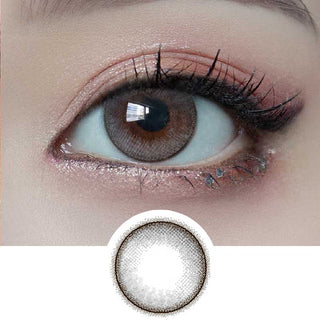 A close-up of an eye wearing Molak 1-Day Dollish Grey (10pk) lenses by Molak, showing a soft gray hue with hazel tones and a dark rim. The eyelid features light pink eyeshadow and long lashes, with the lens design previewed below.
