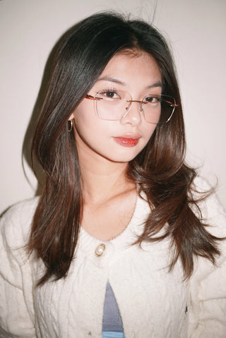 A young woman with long dark hair wears EyeCandys Dusk Rimless Rectangular glasses, a white textured sweater, and a gold hoop earring while posing in front of a plain light background.