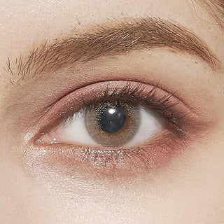 A close-up of a human eye wearing EyeCandys Attitude Glitter Grey contacts by EyeCandys, with soft pink eyelid makeup, a well-groomed eyebrow, and smooth skin illuminated under gentle lighting.