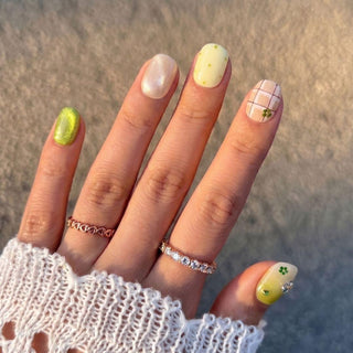 A hand wears EyeCandys Garden Variety Press-On Nails in pastel yellow, green, and white plaid, dot, shimmer, and clover designs—styled with two rings, a white crocheted sleeve, and a glossy salon gel finish.