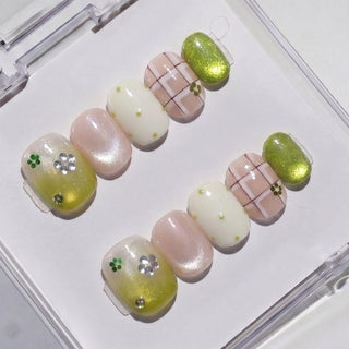 The EyeCandys Garden Variety Press-On Nails set features handmade designs in green, white, and nude with plaid, metallic green, flowers, rhinestones, and dots—neatly arranged in a plastic case for a salon gel look.