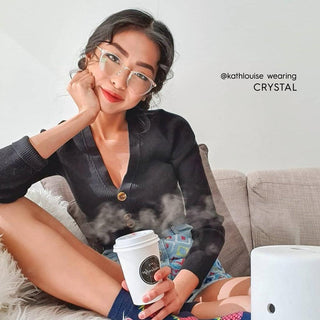 A woman with vintage eyeglasses and a black cardigan sits on a couch, smiling as she holds a steaming cup. Text reads "@kathlouise wearing Infinity Oversized Oval by EyeCandys." A diffuser rests nearby on the sofa.