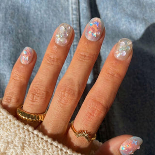 A hand shows off EyeCandys Love Tomorrow Press-On Nails—short, rounded with iridescent, holographic nail art, pearls, and star accents. Gold rings and a cream knit sweater pop against a denim background for a chic salon-style look.