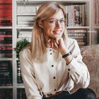A smiling woman with blonde hair wears EyeCandys Luna Round vintage-style titanium glasses and a light button-up shirt, sitting in front of a bookshelf with books in the background, her chin resting on her hand.