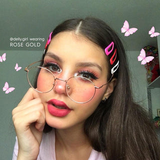 Young woman with long brown hair wears EyeCandys Luna Round titanium glasses in a vintage round frame, plus pink lipstick, bold eye makeup, and colorful hair clips. Pink butterflies and "@delly.girl wearing Luna Round" decorate the image.