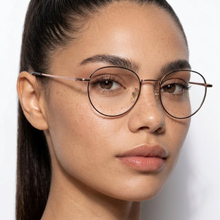 A woman with smooth skin, freckles, and dark hair pulled back wears EyeCandys Luna Round titanium glasses, gazing confidently at the camera against a plain white background.