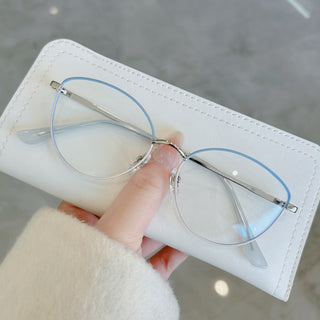 A hand holds a white eyeglass case revealing EyeCandys' Madame Cat Eye glasses—lightweight metal frames with round, thin cat-eye design and a subtle light blue tint on the top edges. The background is blurred gray.