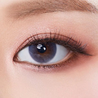 A close-up of a person’s eye with dramatic makeup features Olola Mellows Mystic Grey color contacts by Olola, highlighting long lashes and a well-groomed brow. The striking lenses add extra depth to their look.