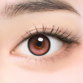 A close-up of an eye with long lashes wearing i-Girl One Pick Balance Peach Bloom Pink contact lens, featuring a dark outer ring. The skin around the eye is smooth and fair.