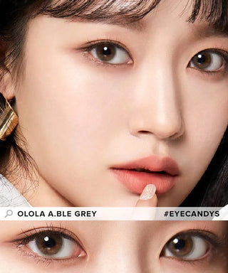 Close-up of a woman with smooth skin, subtle makeup, and Olola Able Grey color contact lens for dark eyes. She holds a finger to her lips and wears a gold earring. Image text: "Olola Able Grey #EYECANDYS".