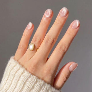 A hand with short, glossy Pink Noise Press-On Nails by EyeCandys is shown wearing a gold ring with a white oval stone and a cream ribbed knit sweater, set against a plain neutral background.