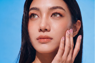 A woman with clear, glowing skin gently touches her cheek, showing off EyeCandys Grapefruit Soda Press-On Nails. Her long dark hair, glossy lips, and natural makeup stand out against the solid light blue background for a fresh look.