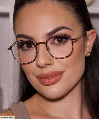 A woman with long brown hair wears EyeCandys Stunner Square glasses, gold earrings, and nude lipstick. She has a nose ring and flawless makeup, smiling slightly while posing indoors.