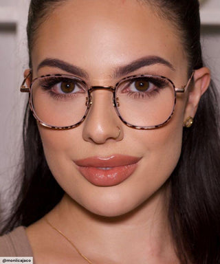 A woman with long dark hair and gold earrings smiles softly. She wears EyeCandys Stunner Square glasses, has full brows, winged eyeliner, nude lipstick, and a nose ring.
