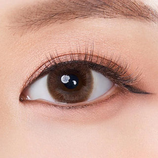 A close-up of a brown eye with long lashes, peach eyeshadow, winged liner, and smooth fair skin wearing Olola Swany Pure Choco contact lens from Olola for a natural enhanced look.