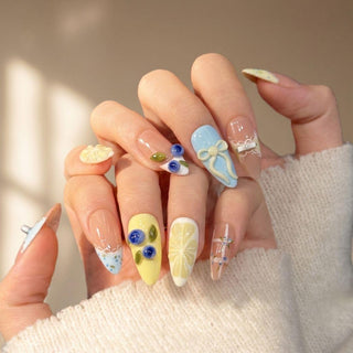 A hand shows off EyeCandys Sweet n Tart Press-On Nails—long, almond-shaped with 3D floral art, pearls, and bows in pastel yellow, blue, and cream—for a salon-style look against a soft beige background.
