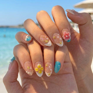 Hands with long, oval EyeCandys Tahiti Feeling Press-On Nails featuring blue polish, white tips, gold stars, 3D yellow and pink flowers, and pearl-like beads held against a beach and ocean backdrop.