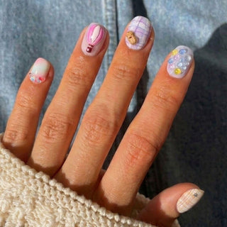 Wearing a cream knit sleeve and denim jacket, a hand displays EyeCandys’ Up Up & Away Press-On Nails—pastel pink hot air balloon, purple bear plaid, gems, mini flowers, and whimsical plaid designs—for reusable style.