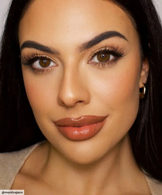 A woman with long dark hair and brown eyes poses in neutral-toned makeup, full brows, defined lashes, contoured cheeks, nude lipstick, gold hoops, a beige top, and EyeCandys Pink Label Vintage Brown lenses for a subtle enlarging effect.
