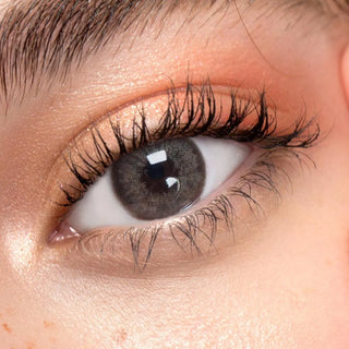 A close-up of an eye with long lashes, peach eyeshadow, and EyeCandys Pink Label Angel Brown contact lens. Smooth skin and a defined brow enhance the look, while the circle lens adds a striking effect.
