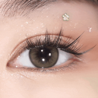 A close-up of an eye with long lashes wearing EyeCandys Pink Label Barcelona Brown contacts. Sparkling makeup accents and a small decorative flower sticker above the brow highlight these comfortable colored lenses.