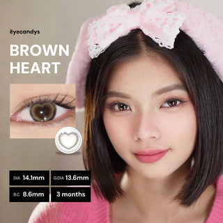 A young woman with straight dark hair, wearing a pink headband and sweater, models EyeCandys Brown Heart contact lenses. A close-up on the left highlights the lens details.