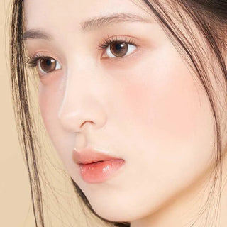 Close-up of a woman with smooth skin, natural makeup, and dark hair wearing Gemhour Demeter 1-Day 13.5 Amber Brown (10pk) contacts for a subtle daily look, set against a beige background. Brand: Gemhour.