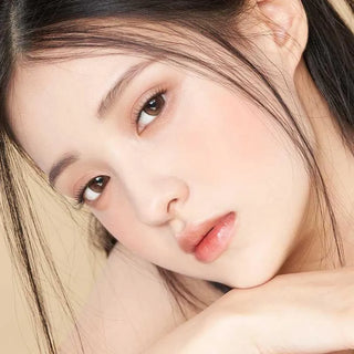 A young woman with clear skin and straight dark hair rests her chin on her hand, wearing Gemhour Demeter 1-Day 13.5 Amber Brown (10pk) contacts that enhance her soft brown eyes against a light beige background.