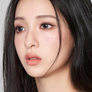 A young woman with long black hair gazes off to the side, wearing Gemhour Demeter 1-Day 13.0 Ocean Blue contacts. She has clear skin, natural makeup, soft pink lips, and a small mole near her nose on her right cheek.