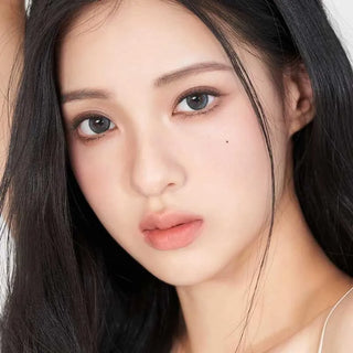 A young woman with long black hair and fair skin wears Gemhour Demeter 1-Day 13.0 Ocean Blue (10pk) contacts, has natural makeup, soft pink lips, a small beauty mark on her right cheek, and poses against a plain light background.