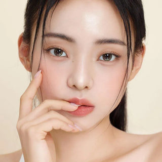 A young woman with clear, glowing skin and natural makeup poses with her fingers gently touching her lips, wearing Gemhour Demeter 1-Day 13.5 Olive Green contacts from Gemhour. She has long black hair and soft pink lips with a calm expression.