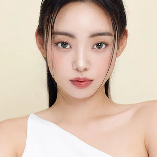 A woman with straight black hair and clear skin wears Gemhour Demeter 1-Day 13.5 Olive Green color contacts. She has natural makeup, a white one-shoulder top, and poses against a light beige background.