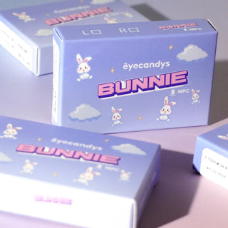 Lavender boxes labeled "EyeCandys Bunnie Milky Ash Grey" with cartoon bunnies, clouds, and stars sit on a matching surface. The pastel packaging features playful retro pixel text, reflecting the glowy design of these EyeCandys lenses.