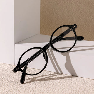 A pair of black EyeCandys Chase Round Oval glasses sits on a white geometric surface, casting gentle shadows against a textured beige backdrop.